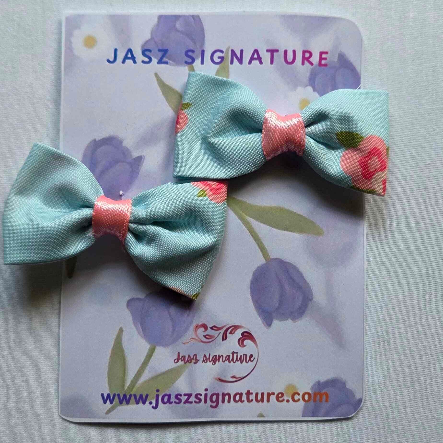 Two light blue hair bows with pink centers on a Jasz Signature card with floral design.