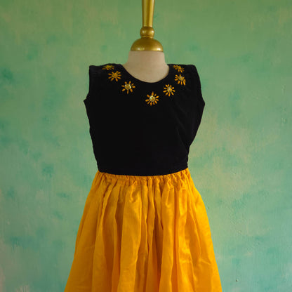 Black top with yellow floral embellishments and yellow skirt on a mannequin against a teal background