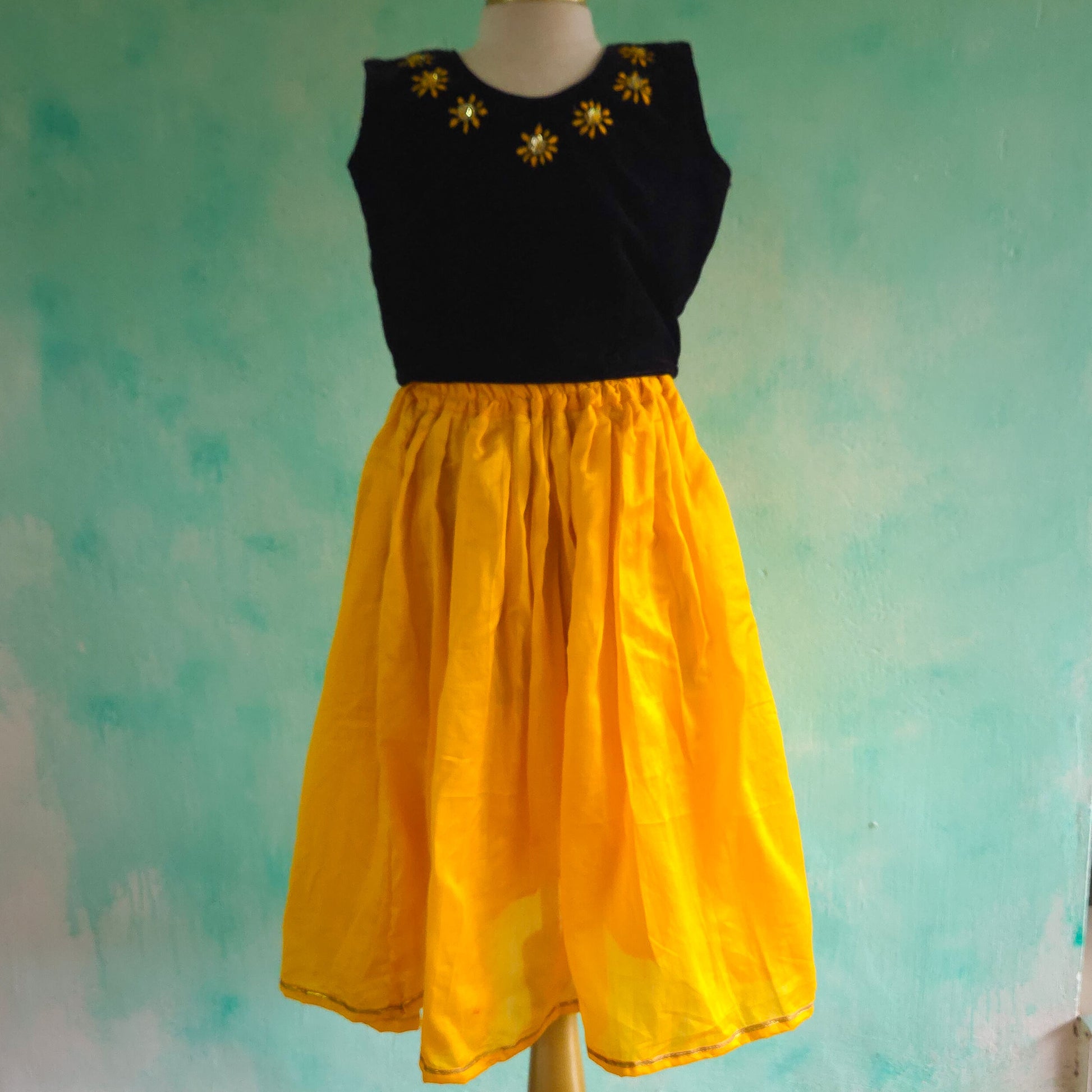Black top with gold embellishments and yellow skirt on a mannequin against a textured teal background