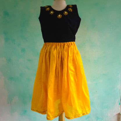 Black top with gold embellishments and yellow skirt on a mannequin against a textured teal background